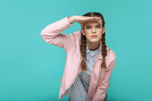 looking too far. portrait of beautiful cute girl standing with makeup and brown pigtail hairstyle in striped light blue shirt pink jacket. indoor, studio shot isolated on blue or green background.